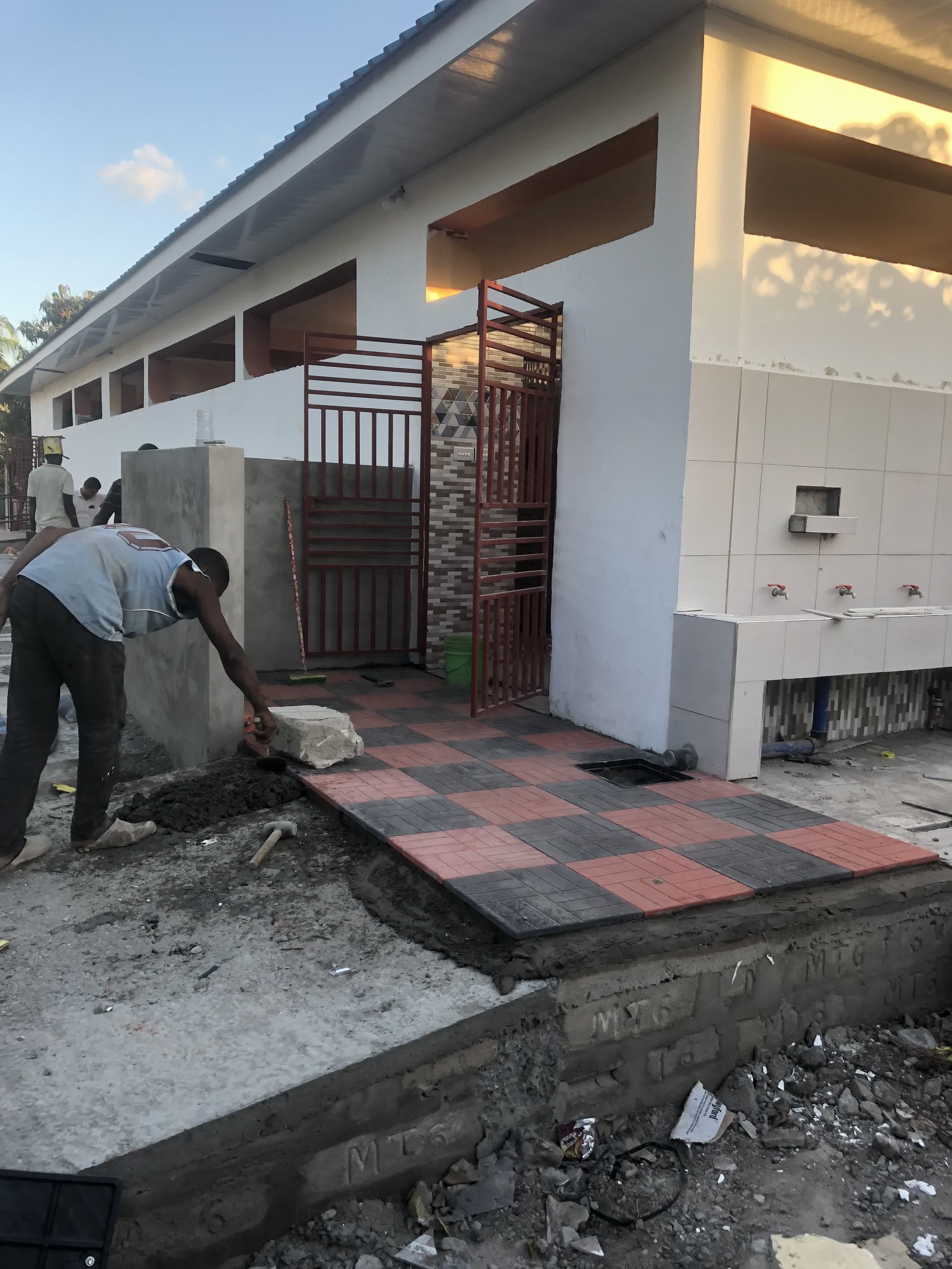 A worker laying patterned tiling beside a metal gate during the finishing phase of a Kikwata project